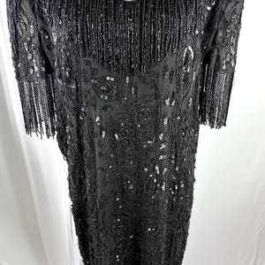 May include: Black beaded dress with a V-neck and short sleeves. The dress has detailed sequin and bead work, with fringe along the hem and sleeves. The dress is displayed on a mannequin.