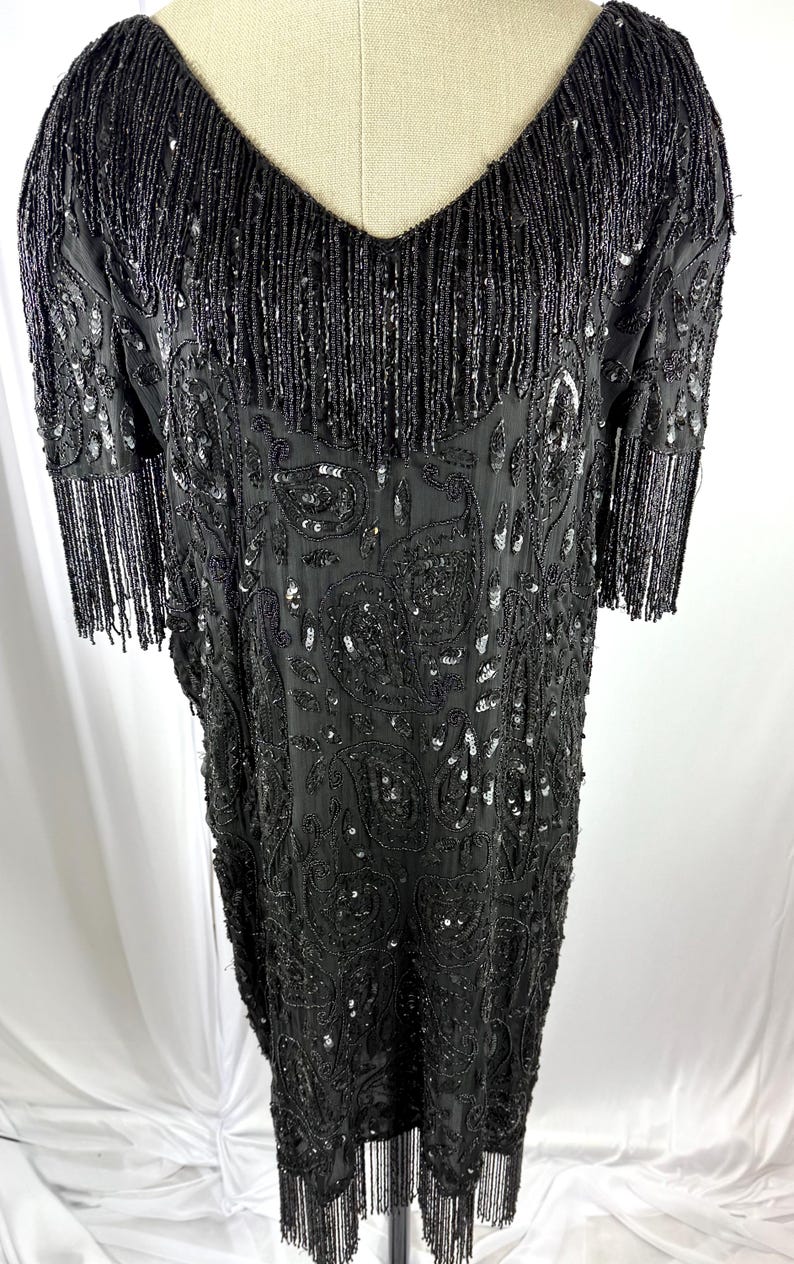 May include: Black beaded dress with a V-neck and short sleeves. The dress has detailed sequin and bead work, with fringe along the hem and sleeves. The dress is displayed on a mannequin.