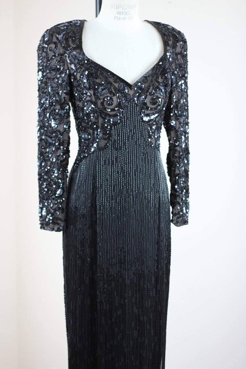 May include: A long-sleeved black sequined dress with a V-neckline. The dress has a fitted bodice and a flowing skirt. The sequins are arranged in a pattern that creates a shimmering effect.
