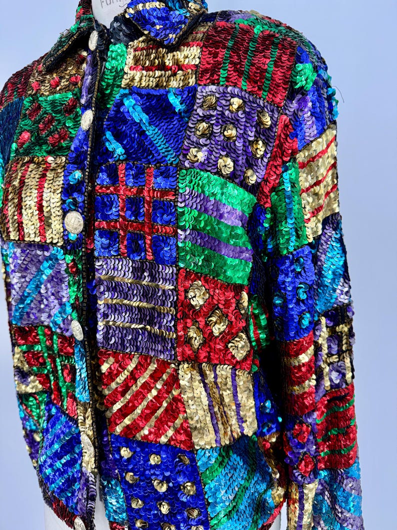 Puede incluir: Una colorida chaqueta de lentejuelas con dise&ntilde;o de patchwork. La chaqueta presenta una variedad de colores, incluyendo rojo, azul, verde, dorado y morado. Las lentejuelas est&aacute;n dispuestas en un patr&oacute;n geom&eacute;trico, creando un aspecto texturizado y llamativo. La chaqueta tiene cuello y botones.