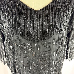 May include: Black beaded dress with a V-neckline and short sleeves. The dress features a paisley pattern with sequins and fringe detailing along the neckline and sleeves. The dress is made of a sheer fabric.