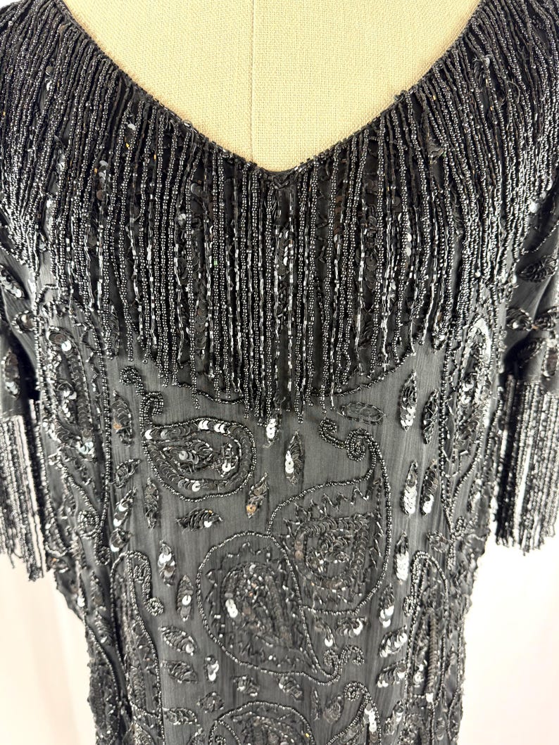 May include: Black beaded dress with a V-neckline and short sleeves. The dress features a paisley pattern with sequins and fringe detailing along the neckline and sleeves. The dress is made of a sheer fabric.