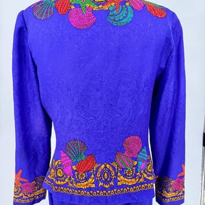 May include: A vibrant blue two-piece suit featuring a seashell design. The jacket and skirt set has a detailed pattern with colorful seashells and starfish, outlined in gold. The suit is displayed on a mannequin.