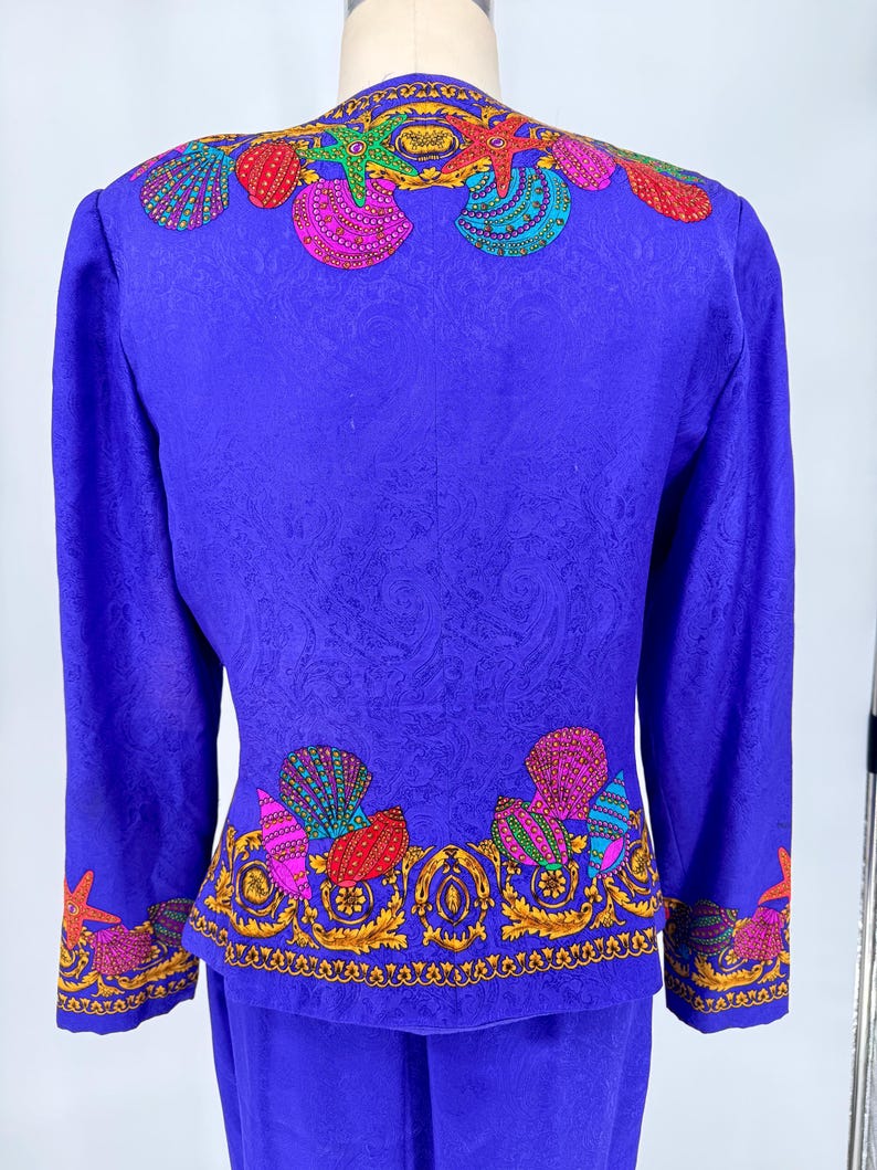 May include: A vibrant blue two-piece suit featuring a seashell design. The jacket and skirt set has a detailed pattern with colorful seashells and starfish, outlined in gold. The suit is displayed on a mannequin.