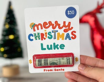 Puede incluir: Una tarjeta blanca con un mensaje "Feliz Navidad Luke" en rojo, amarillo, azul y verde. Un billete de dólar rojo y blanco está metido en una ventana de plástico transparente en la tarjeta. La tarjeta también tiene un círculo azul con el texto "$50" y el texto "De: Santa Claus".