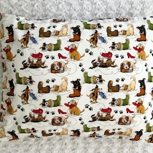 May include: Rectangular pillow with a white background covered in a repeating pattern of playful dogs, boots, shoes, and paw prints. The dogs are in various poses, some holding shoes or boots. The color palette includes brown, red, green, and black.