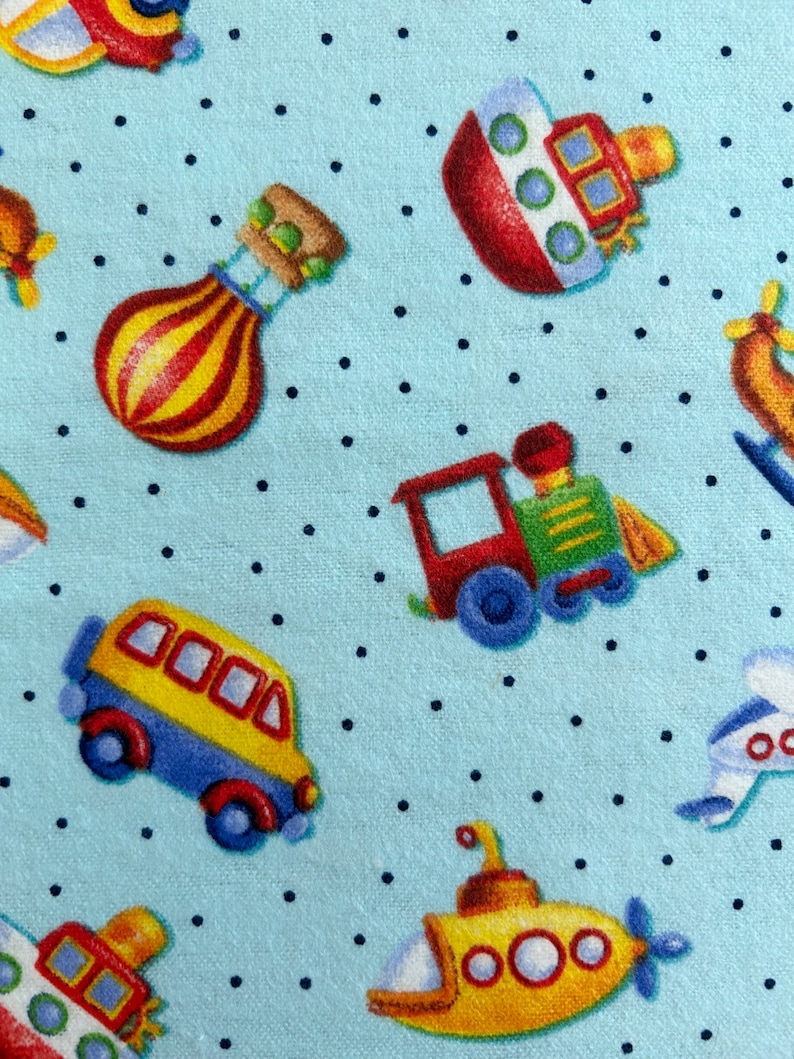 May include: A blue fabric with a white polka dot pattern featuring various colorful cartoon vehicles including a red boat, a yellow submarine, a red train, a yellow school bus, a hot air balloon, and a blue airplane.