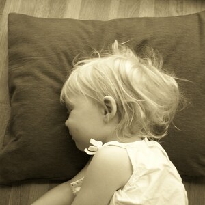 May include: A young child with blonde hair is sleeping on a brown pillow. The child is wearing a white sleeveless shirt.