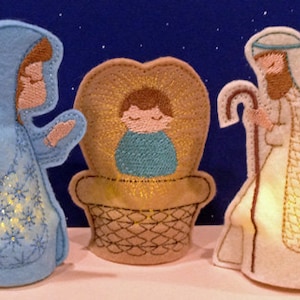 May include: A set of three felt Christmas ornaments depicting the Nativity scene. Mary is wearing a blue robe with white snowflakes, Joseph is wearing a white robe with a gold pattern, and baby Jesus is in a brown manger with a gold halo.