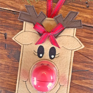 Könnte beinhalten: Ein Filz-Rentier-Anhänger mit einer roten Schleife, braunen Geweih und einer durchsichtigen Plastiknase, die ein rotes Bonbon hält. Der Anhänger hängt an einem rot-grünen Band.
