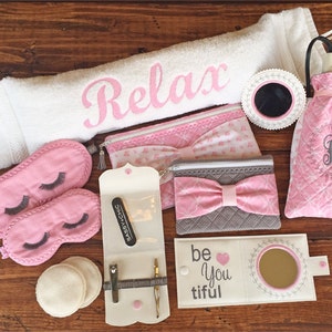 May include: A pink and white spa gift set with a white towel, eye mask, makeup bag, and a small mirror. The towel has the word "Relax" embroidered on it. The makeup bag has a pink bow and the words "be you tiful" on it.
