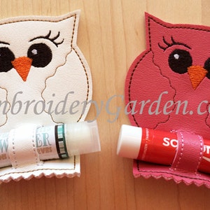 May include: Two owl-shaped lip balm holders. One is white with an orange beak and black eyes, holding a lip balm. The other is pink, also with an orange beak and black eyes, holding a lip balm. The text "EmbroideryGarden.com" is visible.