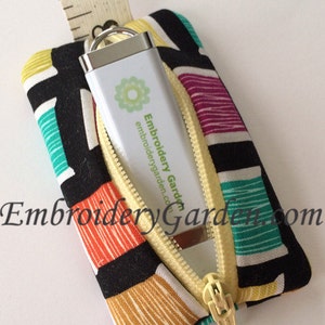 May include: A patterned fabric USB drive case with a yellow zip, containing a white USB drive. The case has a geometric design in black, white, yellow, orange, and teal. The USB drive displays the text "Embroidery Garden" and a logo.
