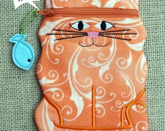 May include: Orange cat-shaped zippered pouch with a white swirl pattern. The pouch has a zipper closure and a small blue fish charm attached to it. The text "in the hoop" is written on a white speech bubble.
