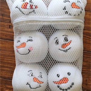 May include: A white mesh laundry bag with six white snowman-shaped balls inside. The snowmen have black eyes, mouths, and buttons, and orange carrot noses.