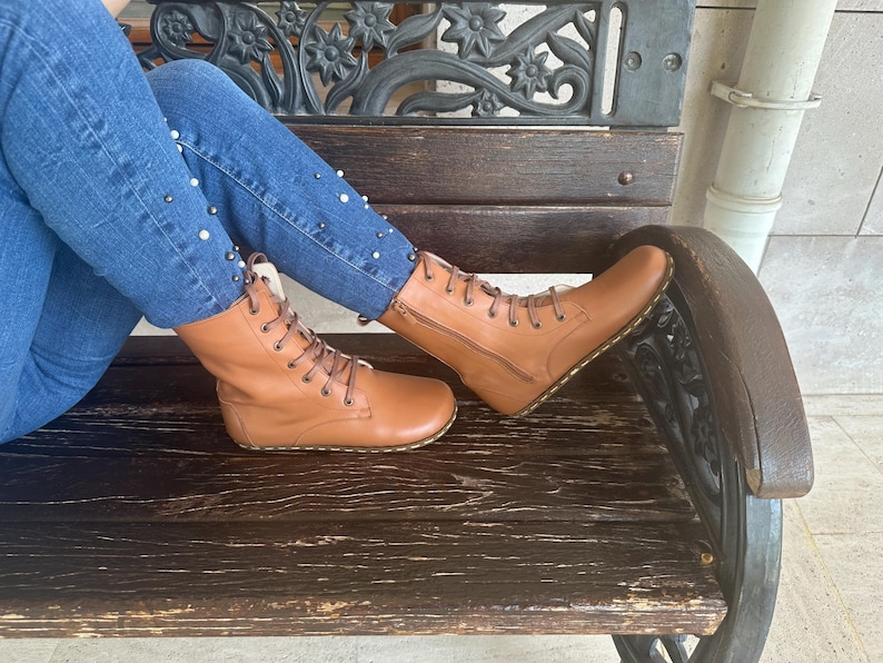 May include: A pair of tan leather boots with laces and zippers. The boots are worn with blue jeans decorated with white pearls. The person is sitting on a dark wooden bench.