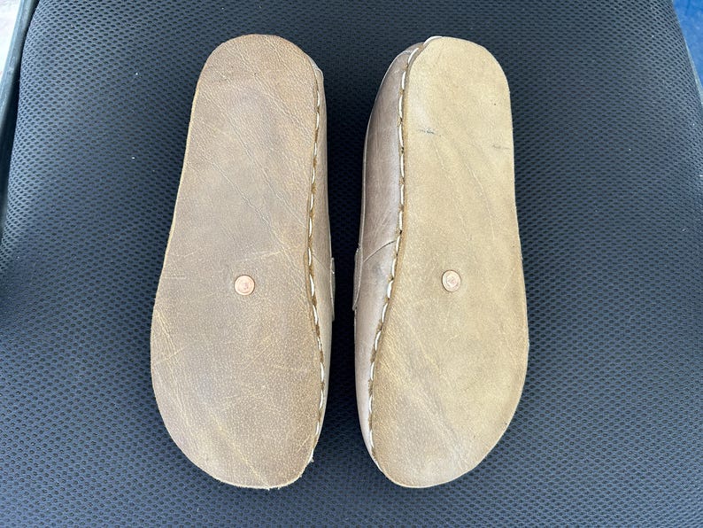 May include: A pair of light brown leather sandals, viewed from the soles. The sandals have a rounded toe design and are stitched with white thread around the edges. A small, circular detail is visible on each sole.