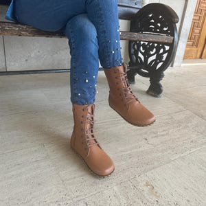 May include: Tan leather lace-up boots with a side zipper. The boots have a flat sole and are worn with blue jeans. The jeans are decorated with small, white pearl-like embellishments. The boots are mid-calf height.