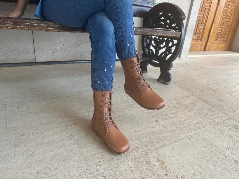 May include: Tan leather lace-up boots with a side zipper. The boots have a flat sole and are worn with blue jeans. The jeans are decorated with small, white pearl-like embellishments. The boots are mid-calf height.