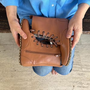 May include: A brown leather handbag shaped like a boot, featuring a zipper and lace-up design. The bag is held by a person wearing a blue shirt and jeans. The stitching is white.