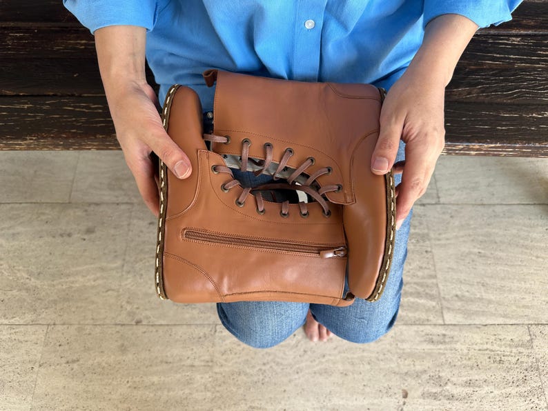 May include: A brown leather handbag shaped like a boot, featuring a zipper and lace-up design. The bag is held by a person wearing a blue shirt and jeans. The stitching is white.