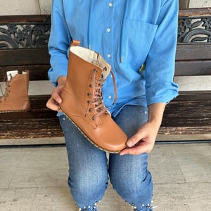 May include: A pair of tan leather lace-up boots. The boots have a round toe, a stitched sole, and a light-colored interior. One boot is held up, while the other sits on a wooden bench. The boots are a light brown color.