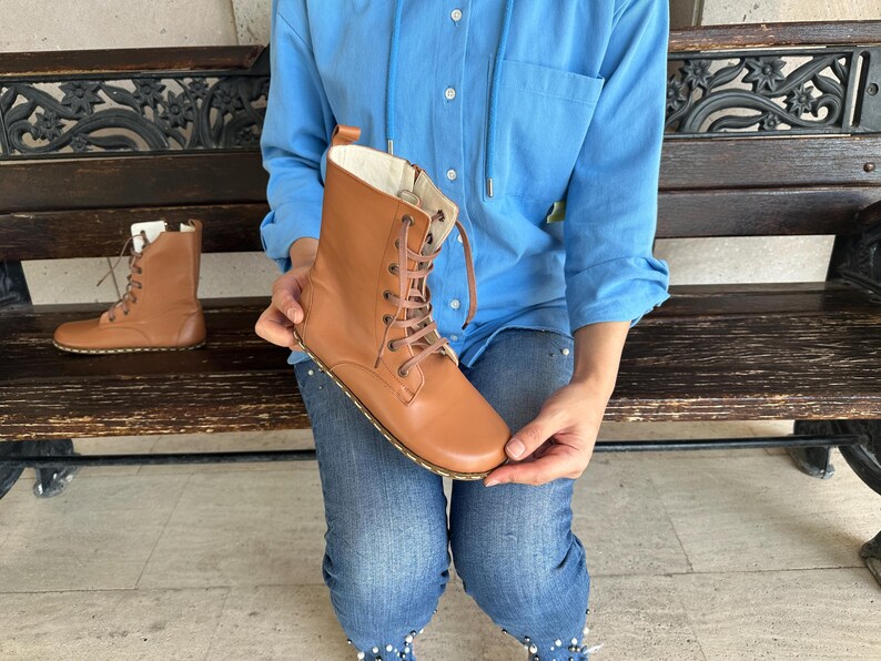 May include: A pair of tan leather lace-up boots. The boots have a round toe, a stitched sole, and a light-colored interior. One boot is held up, while the other sits on a wooden bench. The boots are a light brown color.