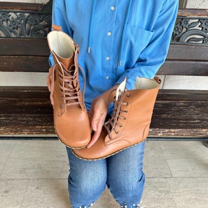 May include: A pair of tan leather boots with laces. The boots have a high-top design and a light-colored interior. The laces are a matching tan color. The boots are held up by a person wearing blue jeans and a blue shirt.