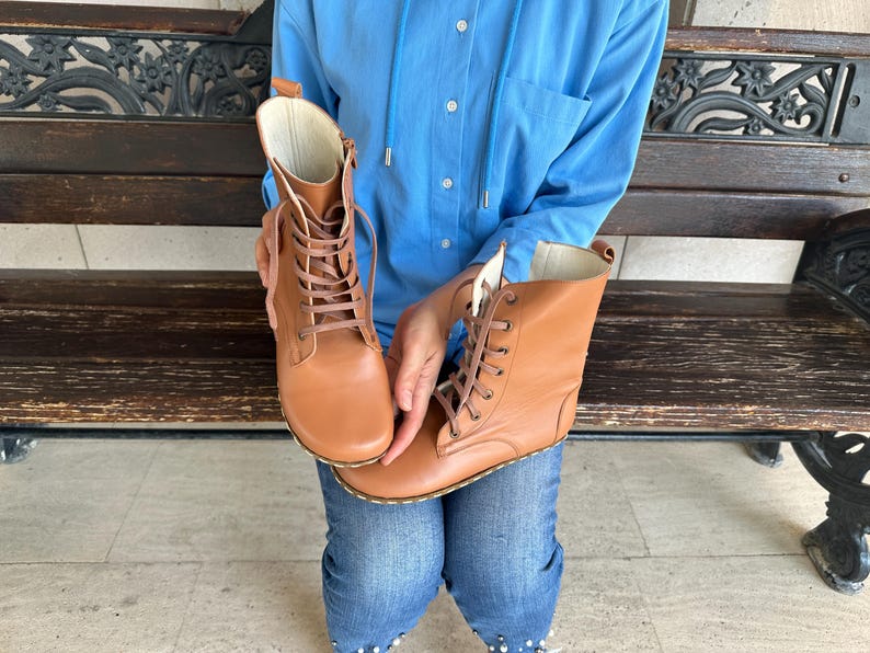 May include: A pair of tan leather boots with laces. The boots have a high-top design and a light-colored interior. The laces are a matching tan color. The boots are held up by a person wearing blue jeans and a blue shirt.