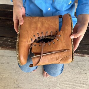 May include: A pair of brown leather boots with laces and zippers. The boots are held by a person wearing a blue shirt and jeans. The boots have a classic design with visible stitching and a worn appearance.