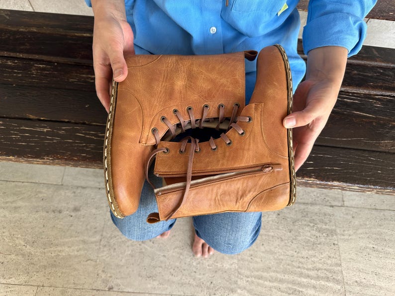 May include: A pair of brown leather boots with laces and zippers. The boots are held by a person wearing a blue shirt and jeans. The boots have a classic design with visible stitching and a worn appearance.