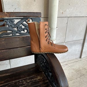 May include: A tan leather boot with a side zipper and lace-up closure. The boot is placed on a wooden bench with decorative metal accents. The boot's design features a flat sole and a mid-calf height.
