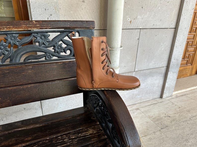 May include: A tan leather boot with a side zipper and lace-up closure. The boot is placed on a wooden bench with decorative metal accents. The boot's design features a flat sole and a mid-calf height.