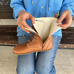 May include: A tan leather boot with a side zipper and lace-up closure. The boot's interior is a light beige color. The boot is being held open, revealing the inside. The sole has a stitched design.