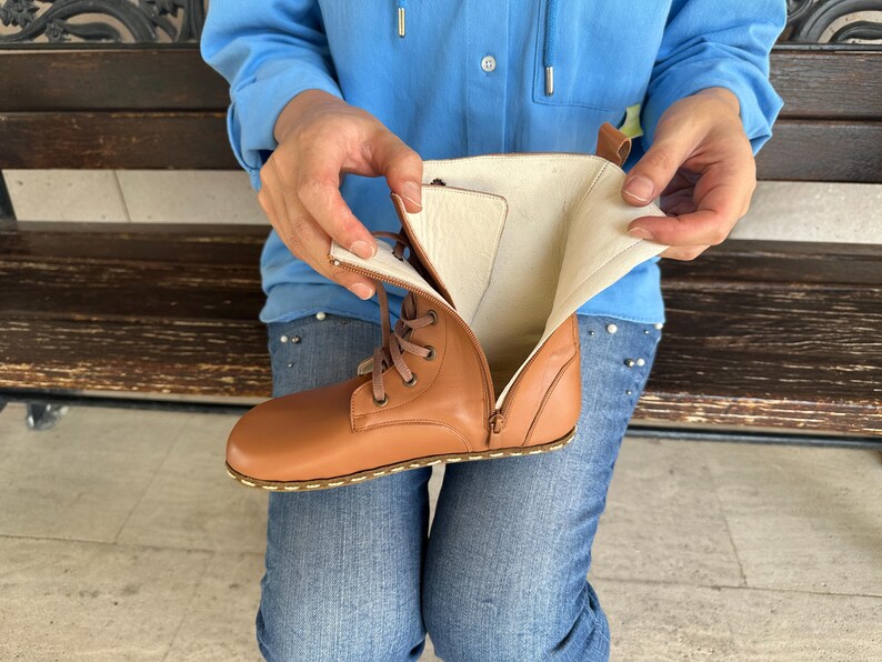 May include: A tan leather boot with a side zipper and lace-up closure. The boot's interior is a light beige color. The boot is being held open, revealing the inside. The sole has a stitched design.