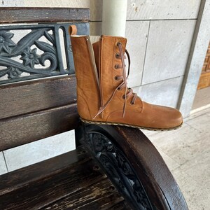 May include: A tan leather boot with a side zipper and lace-up closure. The boot is mid-calf height and has a flat sole. The boot is sitting on a wooden bench with decorative metalwork.
