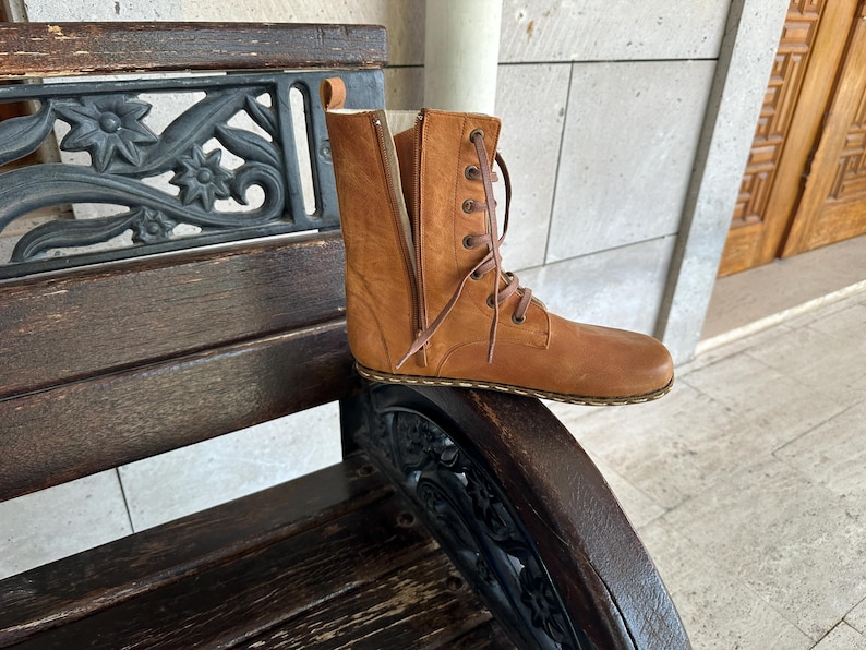 May include: A tan leather boot with a side zipper and lace-up closure. The boot is mid-calf height and has a flat sole. The boot is sitting on a wooden bench with decorative metalwork.