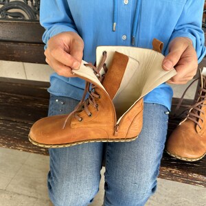 May include: A pair of tan leather boots with laces and zippers. One boot is open, revealing the interior lining. The boots have a classic design and are suitable for various occasions. The boots are shown with a person wearing blue jeans and a blue shirt.