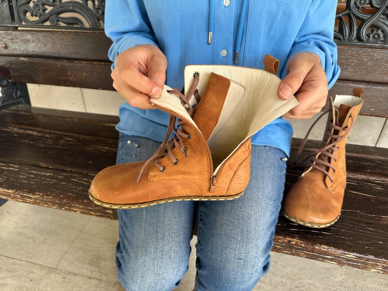May include: A pair of tan leather boots with laces and zippers. One boot is open, revealing the interior lining. The boots have a classic design and are suitable for various occasions. The boots are shown with a person wearing blue jeans and a blue shirt.
