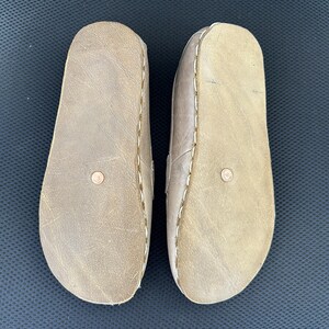 May include: A pair of light brown leather sandals, viewed from the soles. The sandals have a rounded toe design and are stitched with white thread around the edges. A small, circular detail is visible on each sole.