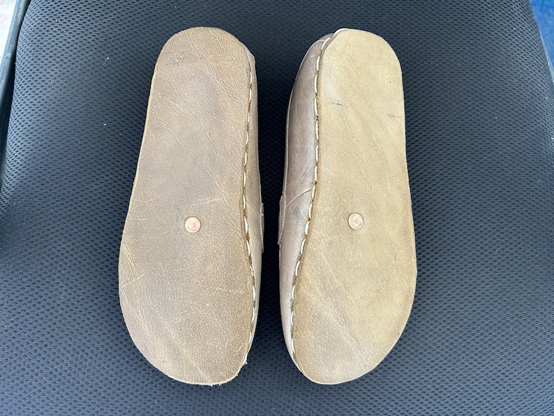 May include: A pair of light brown leather sandals, viewed from the soles. The sandals have a rounded toe design and are stitched with white thread around the edges. A small, circular detail is visible on each sole.