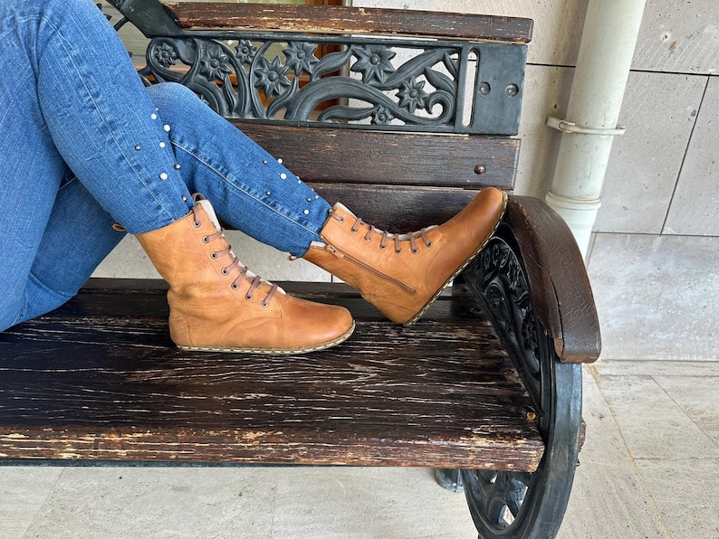 May include: Tan leather boots with laces and a side zipper. The boots have a light-colored interior and are worn with blue jeans. The boots are resting on a wooden bench with decorative metalwork.
