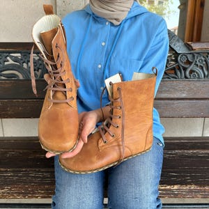 May include: A pair of tan leather boots with brown laces. The boots have a high-top design and are held up by a person. The boots are new and unworn.