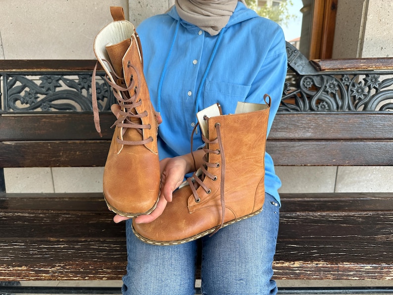 May include: A pair of tan leather boots with brown laces. The boots have a high-top design and are held up by a person. The boots are new and unworn.