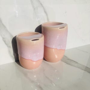 Könnte beinhalten: Zwei Keramikbecher mit einem rosa-weißen Ombre-Glaze. Die Becher haben eine abgerundete Form und eine Lippe zum einfachen Trinken.