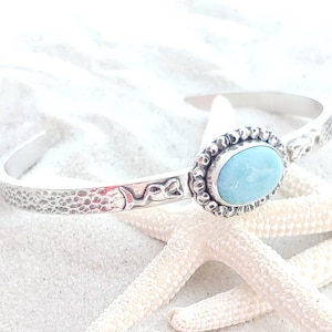 May include: A silver cuff bracelet with a light blue oval gemstone set in a textured bezel. The bracelet has a hammered texture and a decorative design on the sides. The bracelet is displayed on a white surface with starfish.