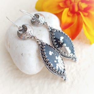 May include: A pair of silver-toned earrings with a dark, speckled, oval-shaped stone. The earrings have a decorative, ornate design with a crescent moon shape at the top and a detailed border around the stone.