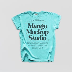 May include: A turquoise t-shirt with rolled sleeves. The shirt displays the text "Mango Mockup Studio" in a bold, black font. Additional text includes "Your Design Here", "Real Product Mockups", "Comfort Colors 1717", and "Chalky Mint".
