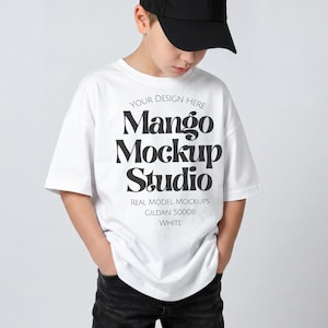 May include: A white t-shirt featuring the text "Mango Mockup Studio" in black, worn by a person wearing a black baseball cap and dark jeans. The shirt also has the text "Your Design Here", "Real Model Mockups", "Gildan 5000B", and "White".