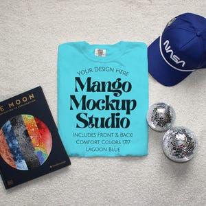 May include: A mockup scene featuring a turquoise t-shirt with the text "Mango Mockup Studio" and a blue NASA baseball cap. Two small disco balls and a book titled "The Moon" are also present.